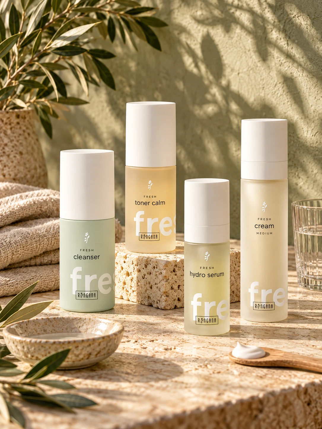 Composición editorial de productos RINGANA FRESH cleanser, toner calm, hydro serum y cream medium sobre piedra natural, con lino, cerámica y hojas de olivo.